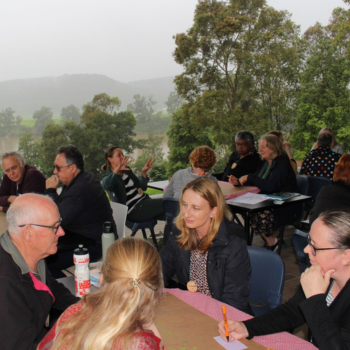 Groups of people seated at outdoor tables engaged in discussion, with a misty forested landscape in the background.